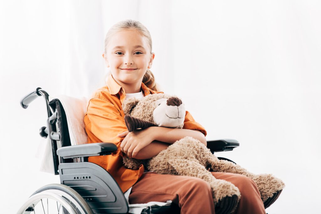 smiling kid holding teddy bear and sitting on wheelchair